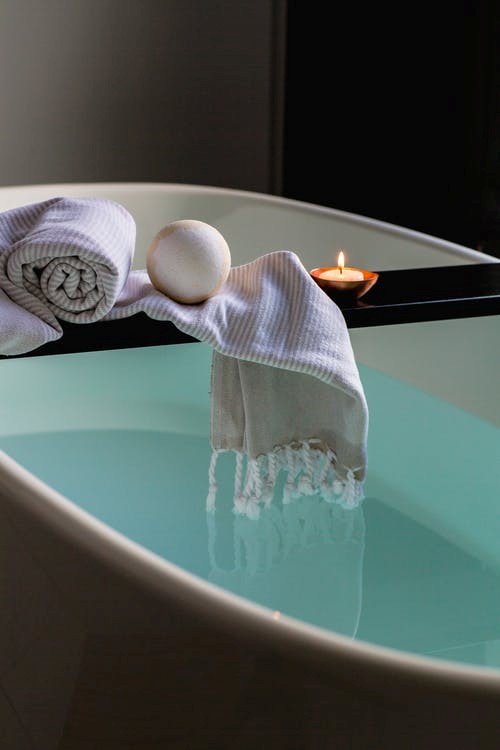 A spa-like setup of a bathtub filled with warm water with a scented candle and towels placed on a panel for a luxury bath.